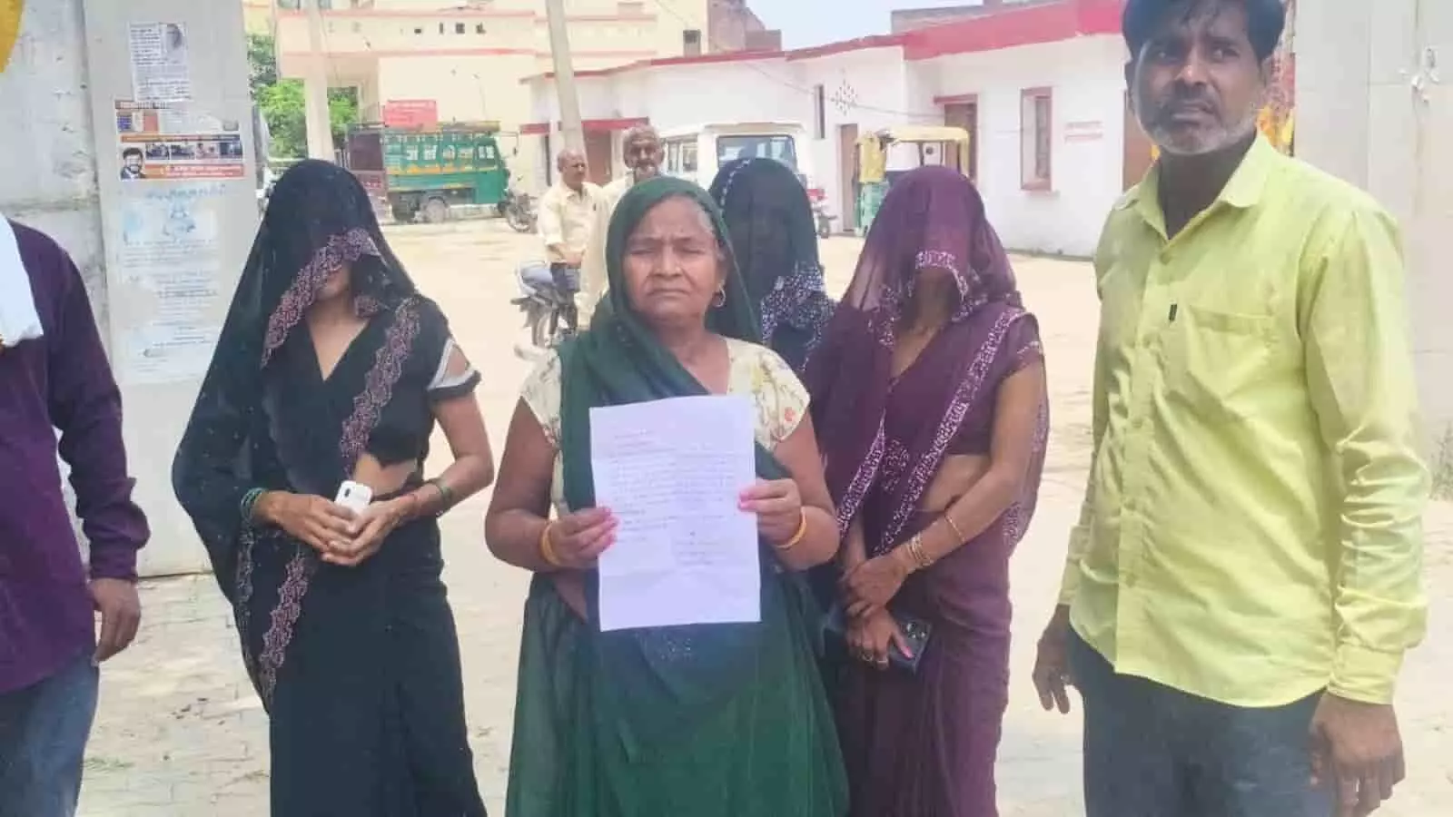 तस्वीर में हाथ में शिकायत पत्र लिए एक महिला और अन्य लोग खड़े दिखाई दे रहे हैं।
