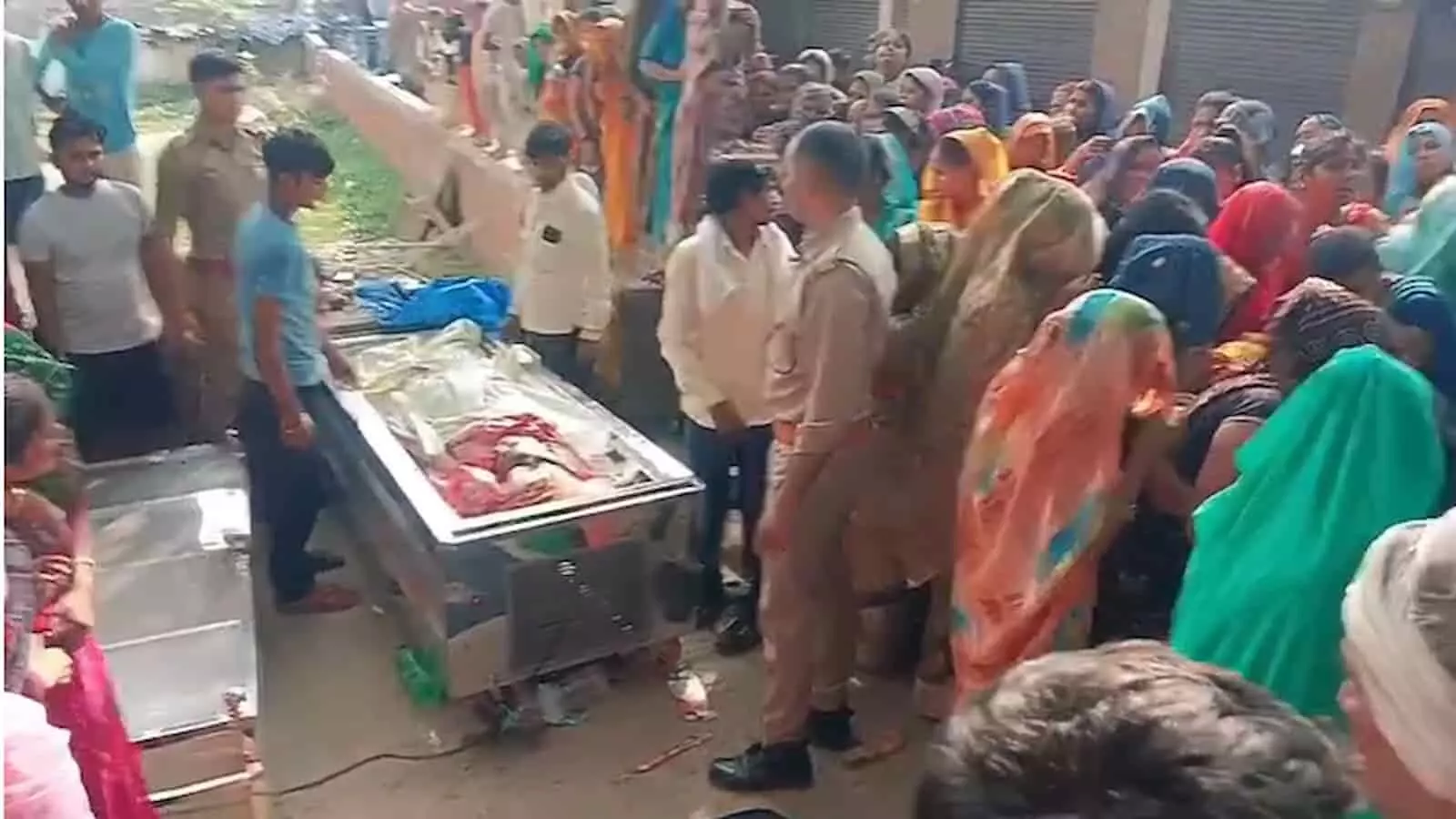 घटना स्थल पर मौजूद भीड़ और पुलिसकर्मी, जहाँ सड़क किनारे शवों का फ्रीजर रखा है।