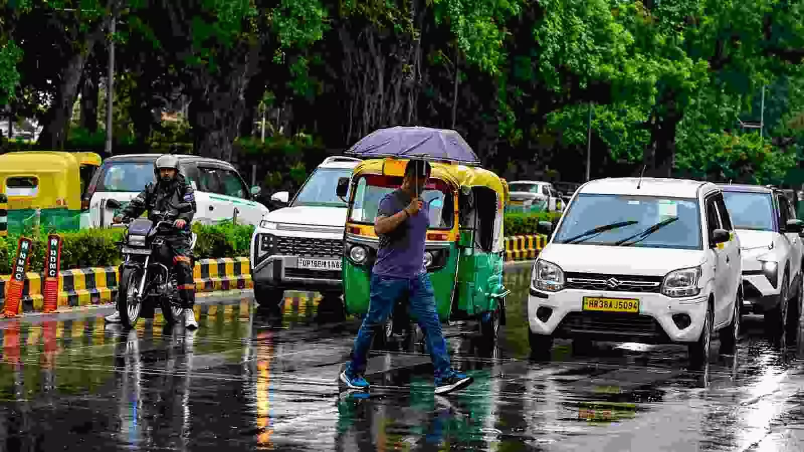 दिल्ली की सड़क पर छतरी लेकर चलते व्यक्ति और वाहनों की आवाजाही का दृश्य।