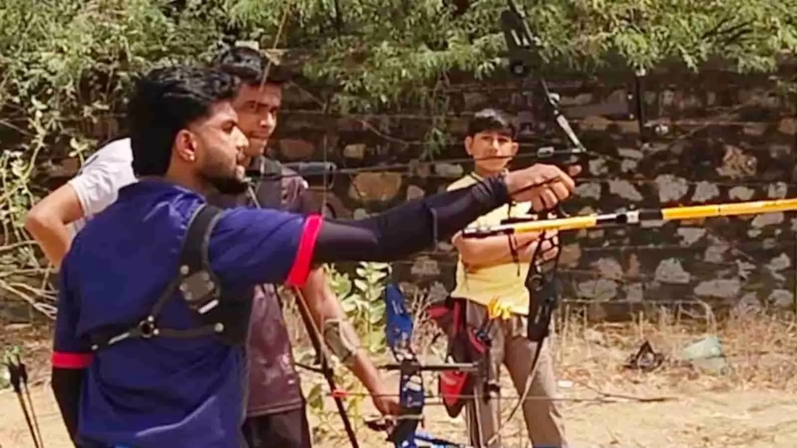 मैदान पर दांतों से धनुष खींचकर निशाना लगाते तीरंदाज गोविंद माली और खड़े अन्य खिलाड़ी। मैदान पर दांतों से धनुष खींचकर निशाना लगाते तीरंदाज गोविंद माली और खड़े अन्य खिलाड़ी।