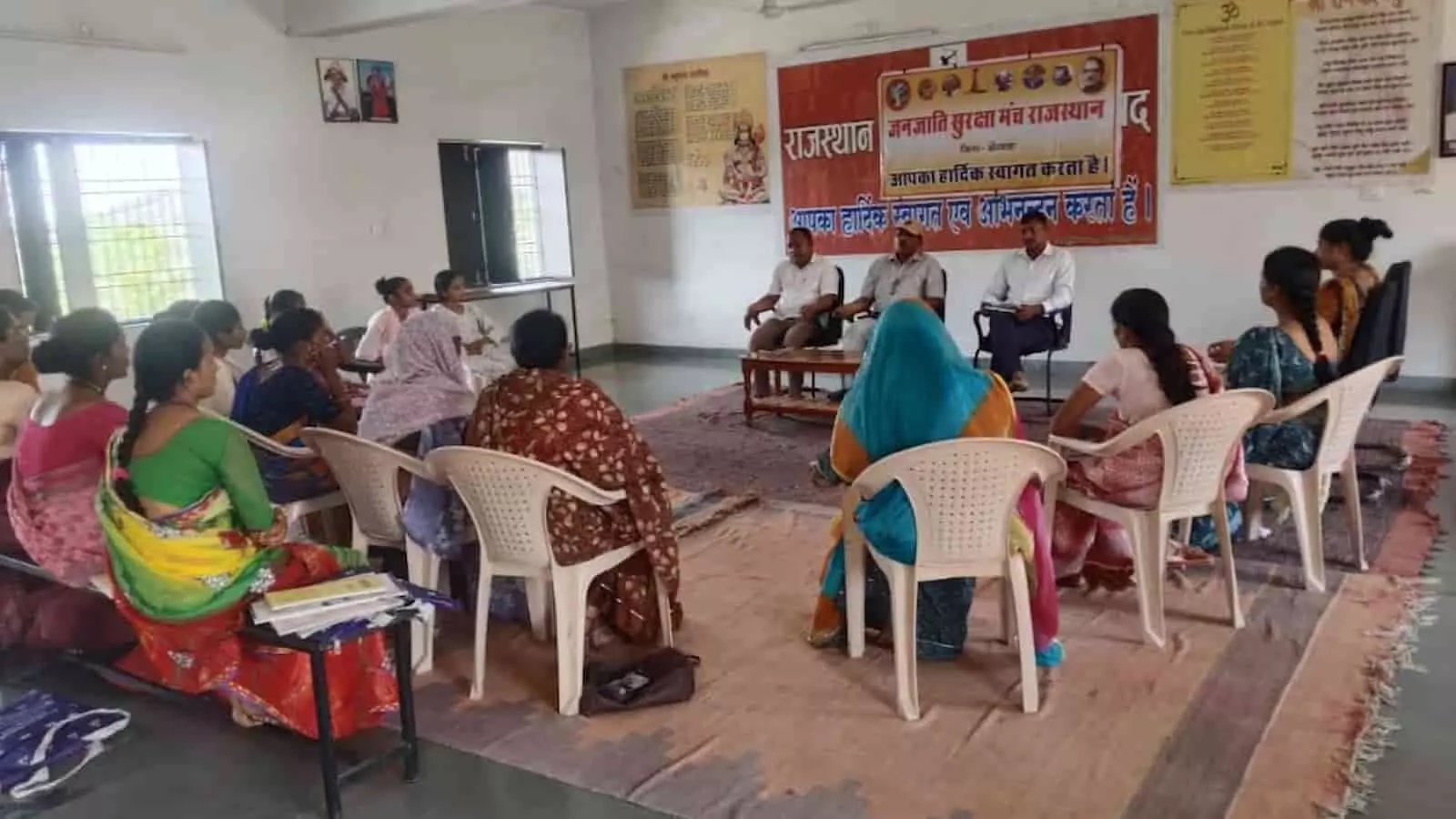 तस्वीर में दिख रही एक इनडोर बैठक जहां महिलाएं और पुरुष कुर्सियों पर बैठकर चर्चा कर रहे हैं।