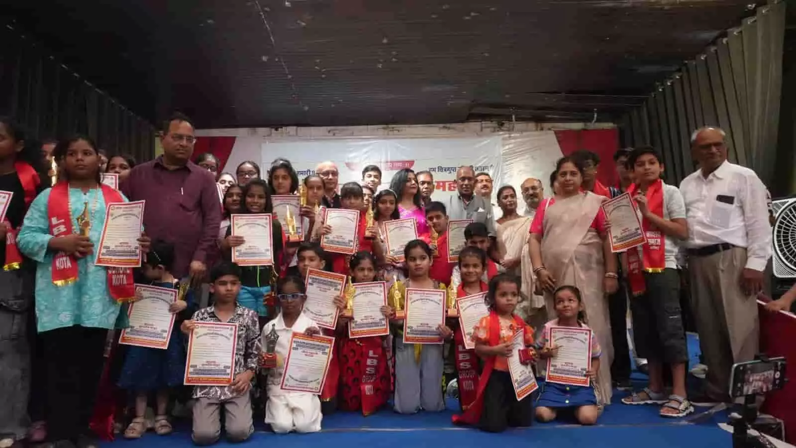 मंच पर ट्रॉफी और प्रमाणपत्र थामे विद्यार्थी तथा अतिथिगण कोटा के सम्मान समारोह में।