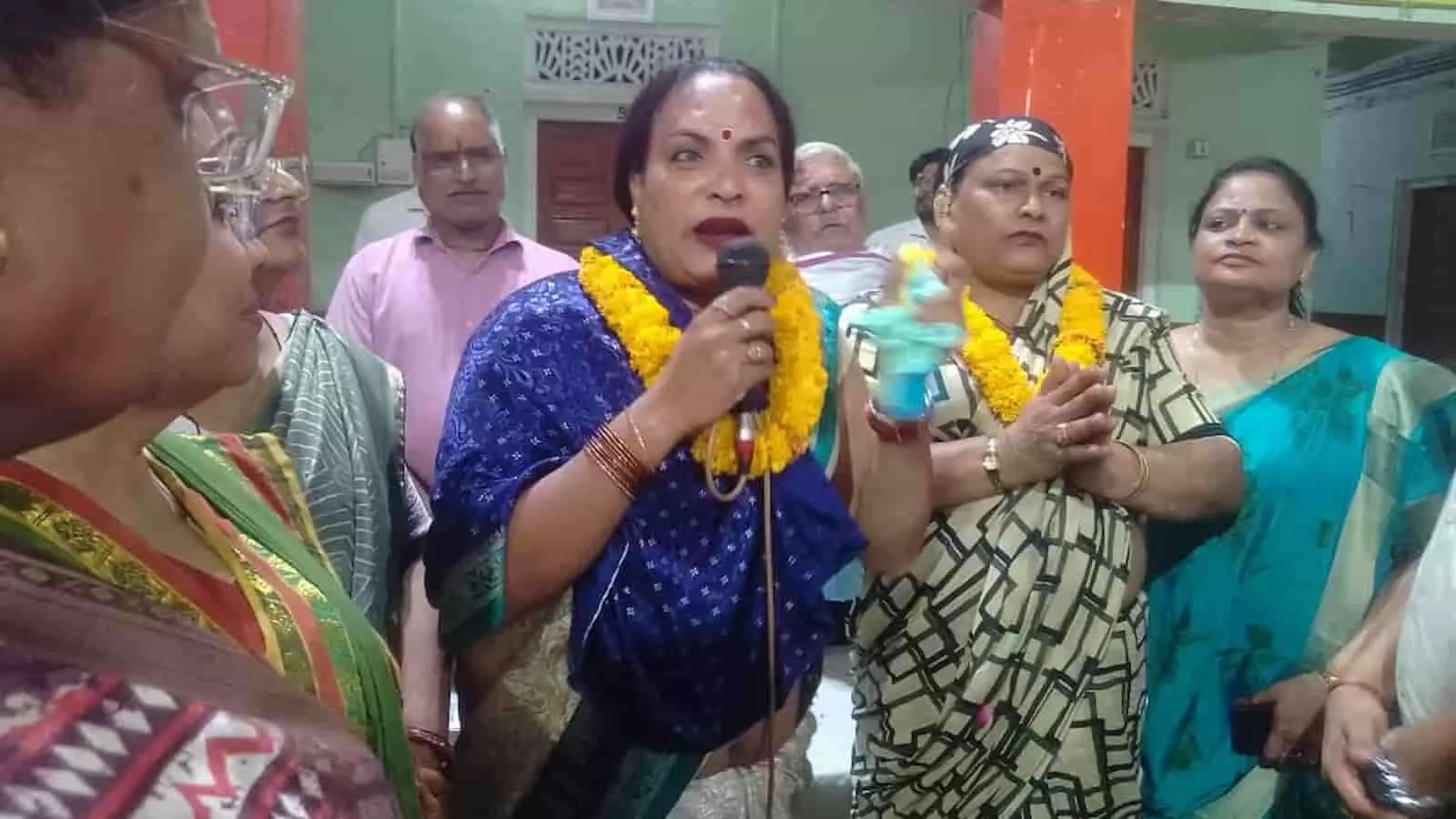 तस्वीर में एक महिला प्रतिनिधि माइक पर बोल रही हैं और अन्य लोग उनके पास खड़े हैं।