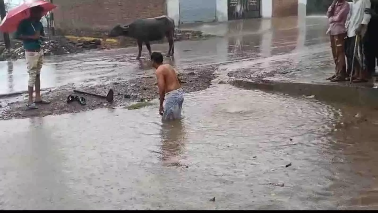 पानी से भरी ग्रामीण सड़क पर खड़ा एक व्यक्ति और खड़े हुए अन्य लोग।