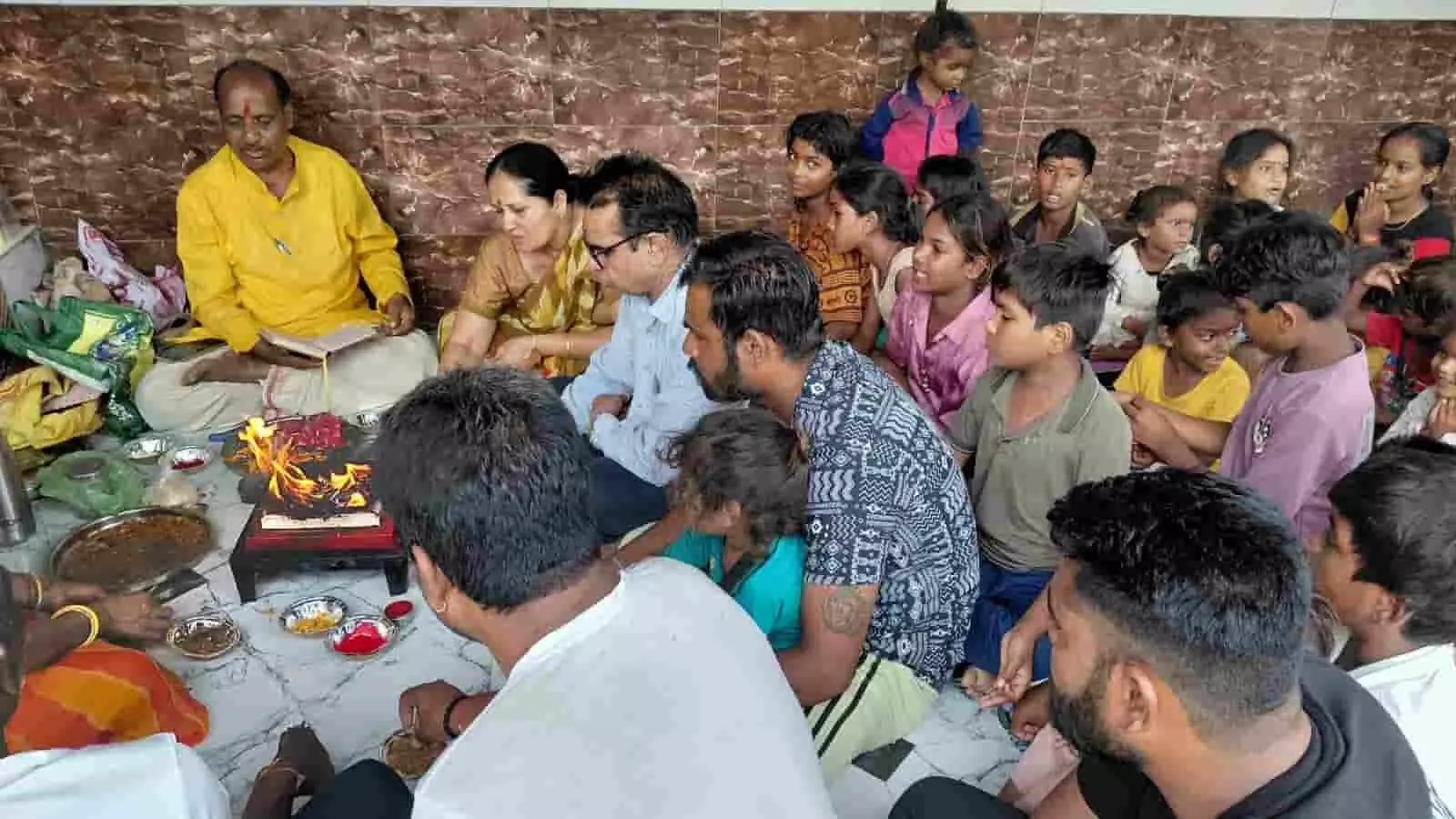 तस्वीर में फर्श पर बैठकर हवन कुंड के पास धार्मिक अनुष्ठान में शामिल होते लोग और बच्चे। तस्वीर में फर्श पर बैठकर हवन कुंड के पास धार्मिक अनुष्ठान में शामिल होते लोग और बच्चे।