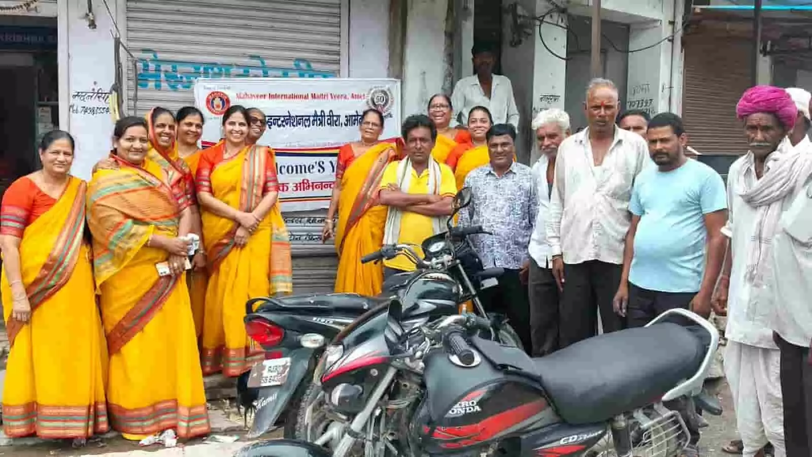 तस्वीर में सड़क सुरक्षा जागरूकता बैनर के सामने महिलाएं और पुरुष मोटरसाइकिल के पास खड़े हैं।