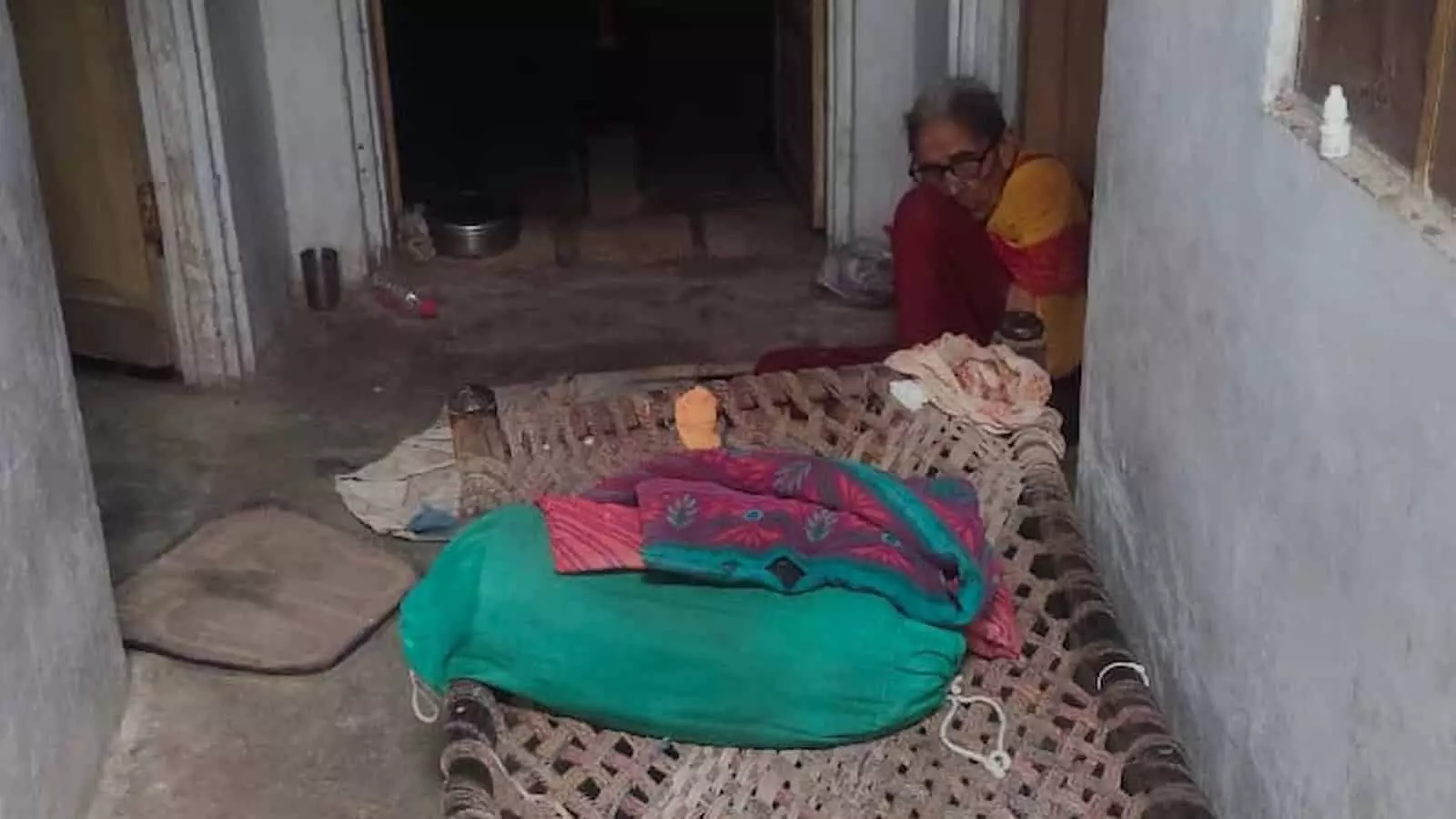 एक पुराने मकान के बरामदे में खाट के पास जमीन पर बैठी बुजुर्ग महिला