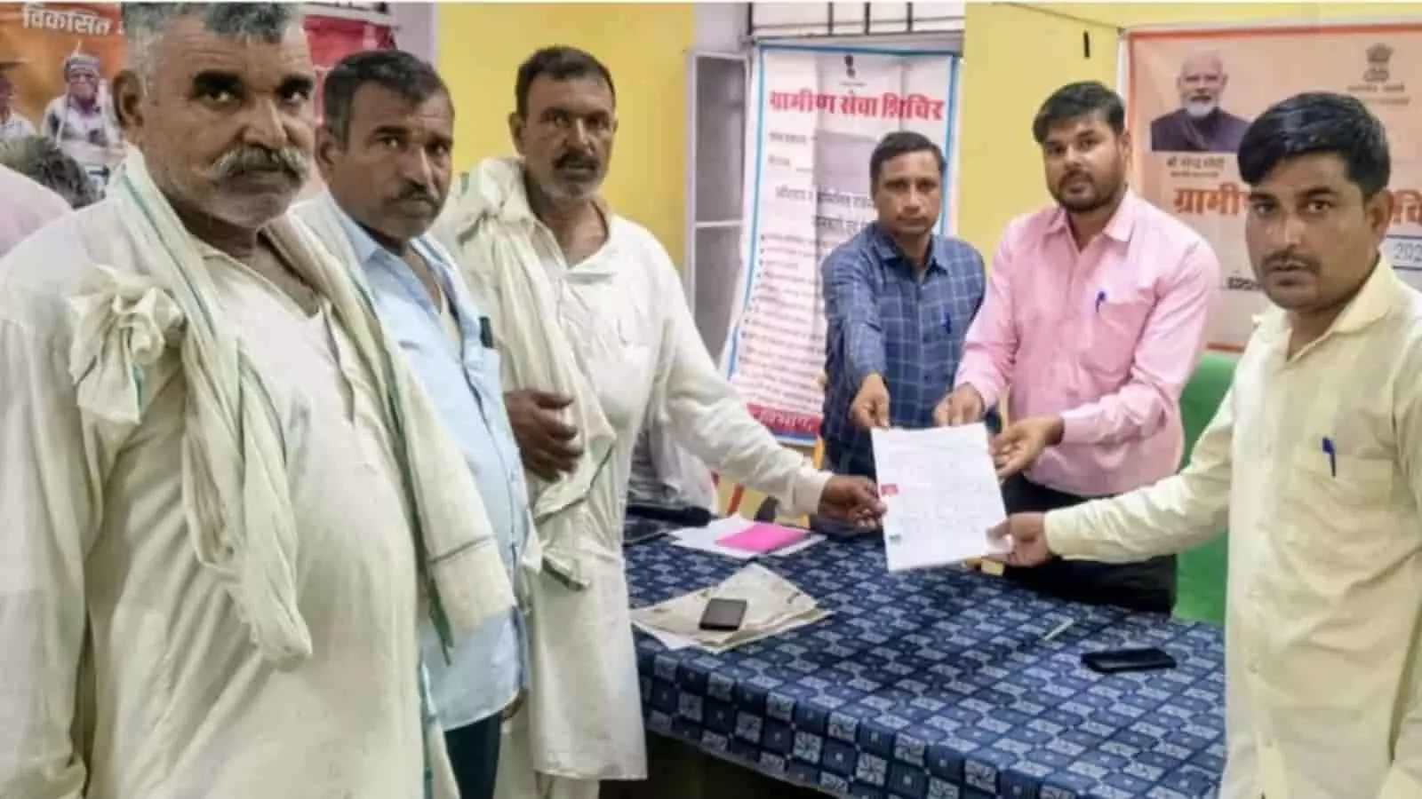 तस्वीर में ग्रामीण सेवा शिविर में अधिकारी और ग्रामीण टेबल के पास दस्तावेज पकड़े हुए हैं। तस्वीर में ग्रामीण सेवा शिविर में अधिकारी और ग्रामीण टेबल के पास दस्तावेज पकड़े हुए हैं।
