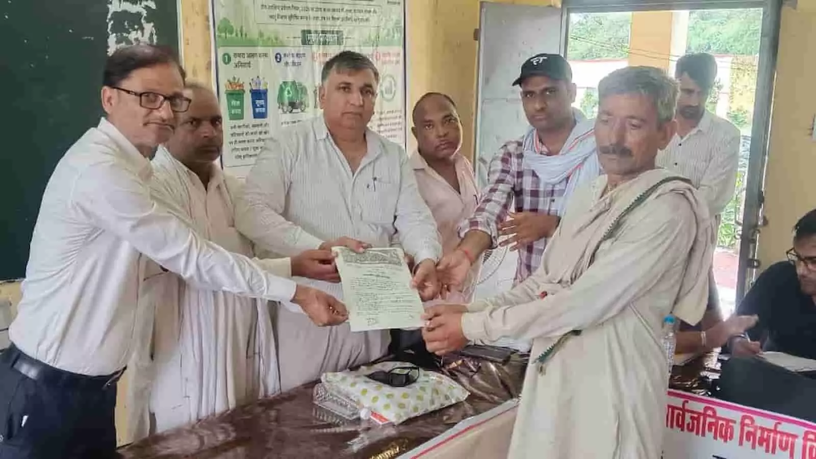 तस्वीर में अधिकारी ग्रामीण सेवा शिविर के दौरान एक ग्रामीण व्यक्ति को आवासीय दस्तावेज सौंपते हुए दिख रहे हैं।