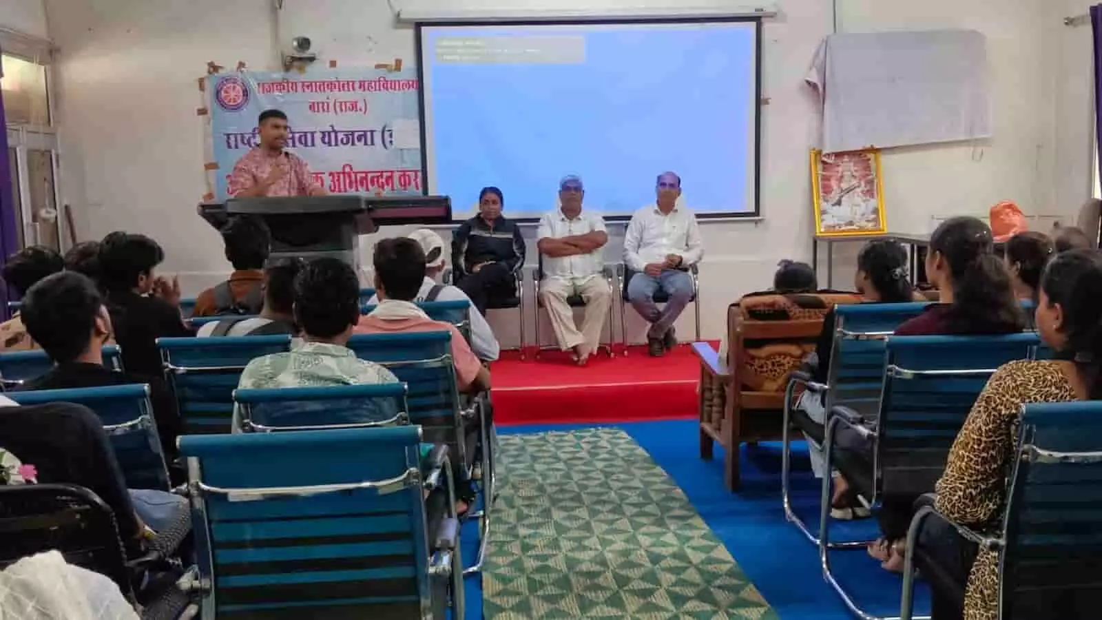 व्याख्यान कक्ष में पोडियम से विद्यार्थियों को संबोधित करते वक्ता और कुर्सियों पर बैठे श्रोता। व्याख्यान कक्ष में पोडियम से विद्यार्थियों को संबोधित करते वक्ता और कुर्सियों पर बैठे श्रोता।