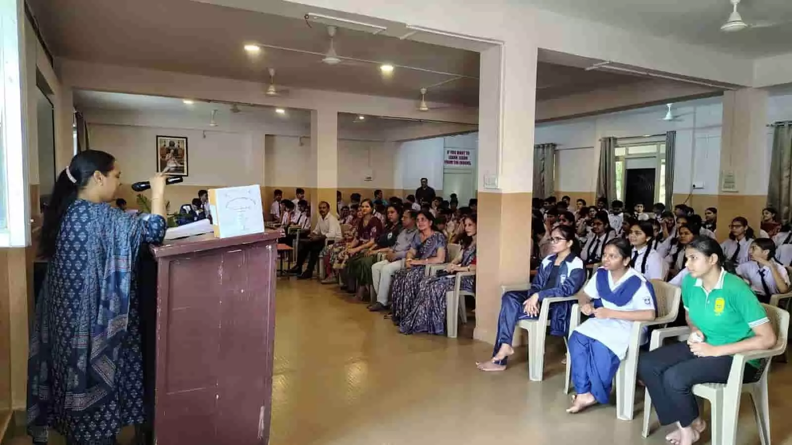 स्कूल के हॉल में मंच से भाषण देती महिला और सामने कुर्सियों पर बैठे विद्यार्थी। स्कूल के हॉल में मंच से भाषण देती महिला और सामने कुर्सियों पर बैठे विद्यार्थी।
