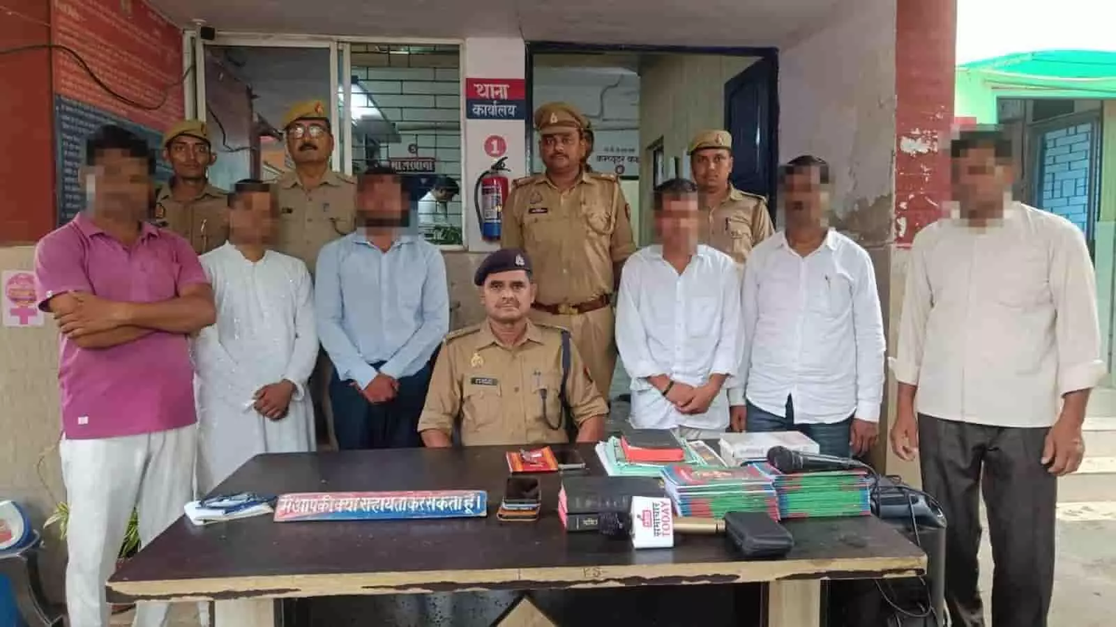 पुलिस स्टेशन में मेज के पास खड़े छह आरोपी और पीछे खड़े पुलिस अधिकारी। पुलिस स्टेशन में मेज के पास खड़े छह आरोपी और पीछे खड़े पुलिस अधिकारी।