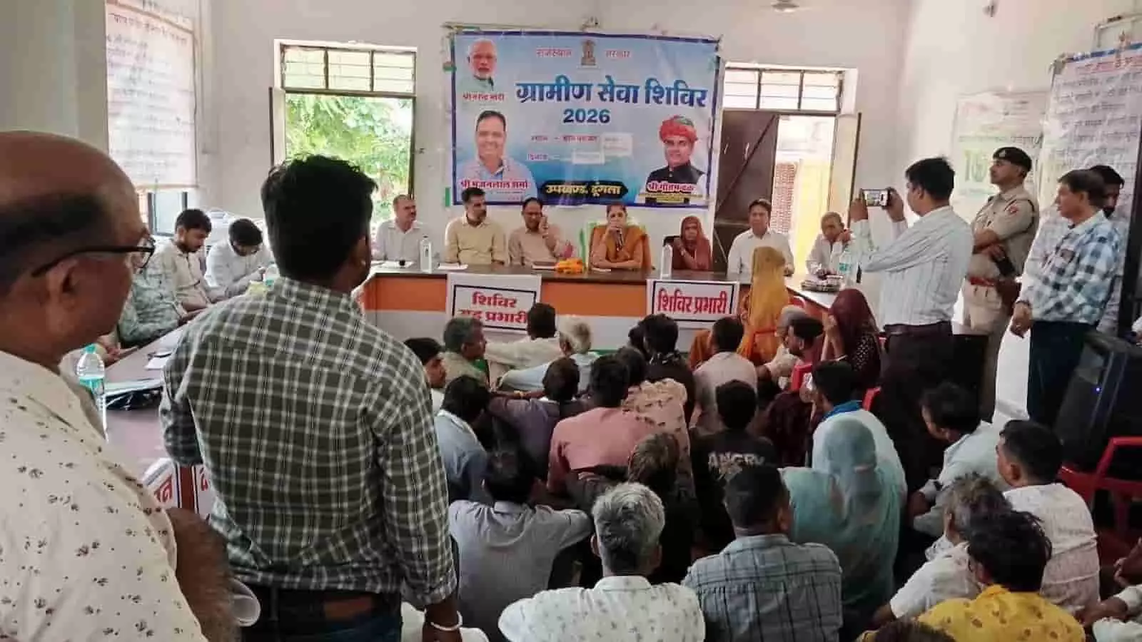 तस्वीर में ग्रामीण सेवा शिविर के दौरान अधिकारी मंच पर बैठे हैं और सामने ग्रामीण उपस्थित हैं। तस्वीर में ग्रामीण सेवा शिविर के दौरान अधिकारी मंच पर बैठे हैं और सामने ग्रामीण उपस्थित हैं।