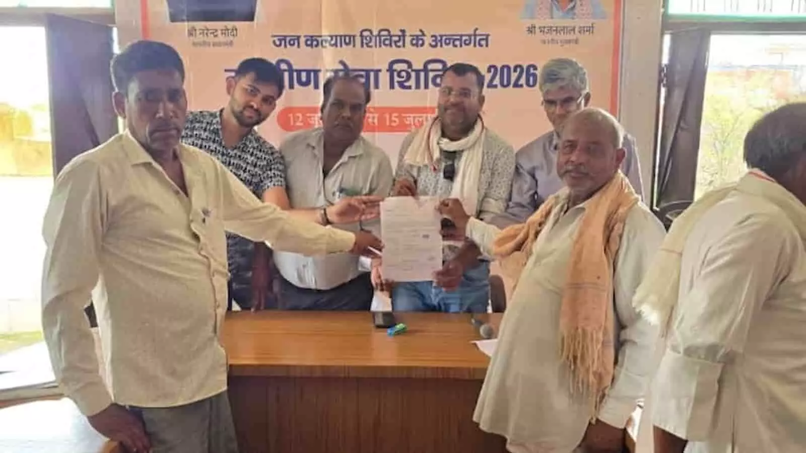 ग्रामीण सेवा शिविर के बैनर तले मेज के पास खड़े होकर सरकारी दस्तावेज दिखाते हुए लोग। ग्रामीण सेवा शिविर के बैनर तले मेज के पास खड़े होकर सरकारी दस्तावेज दिखाते हुए लोग।