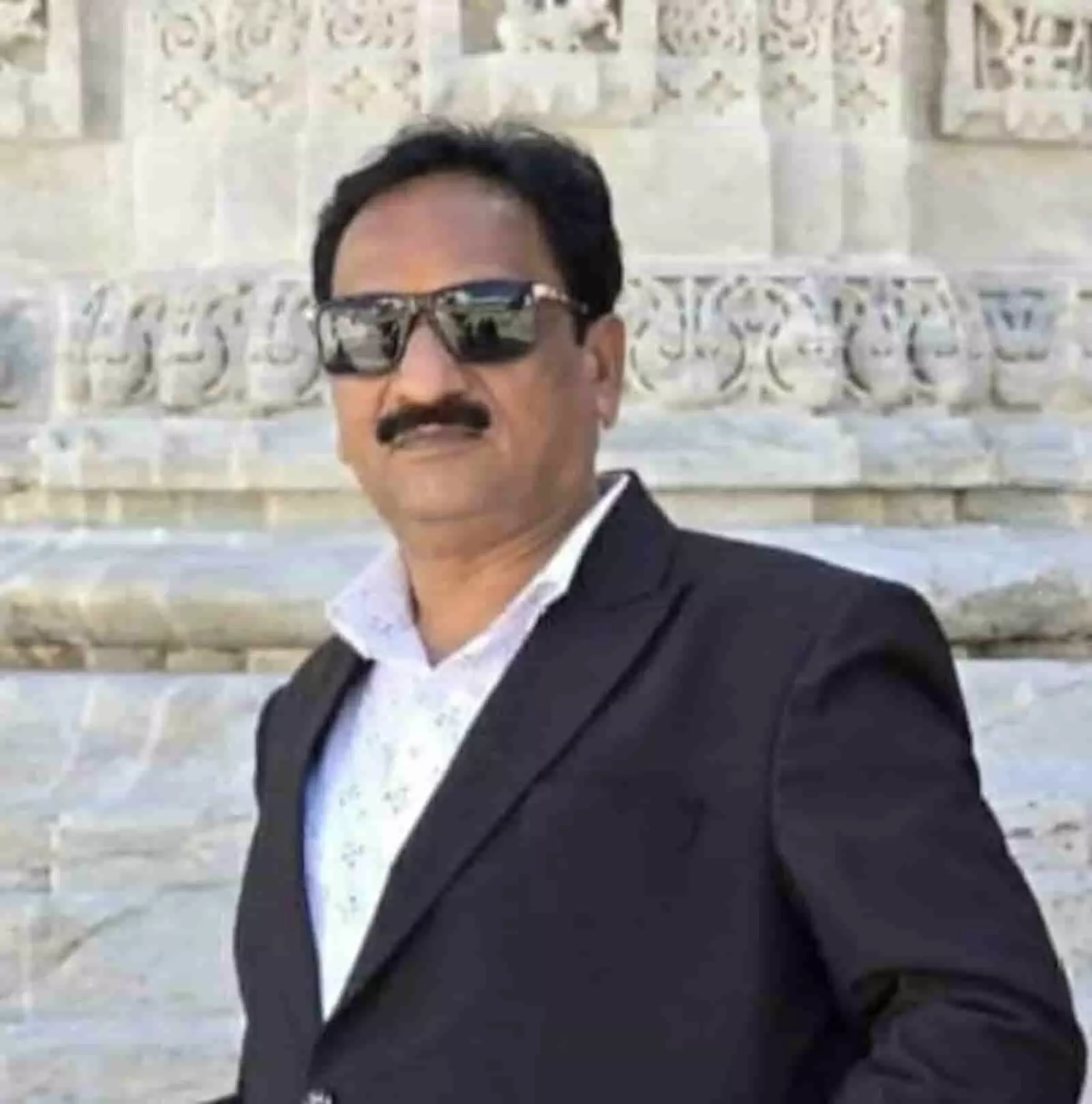 सफेद नक्काशीदार दीवार के सामने काले ब्लेज़र और चश्मे में खड़ा एक व्यक्ति। सफेद नक्काशीदार दीवार के सामने काले ब्लेज़र और चश्मे में खड़ा एक व्यक्ति।