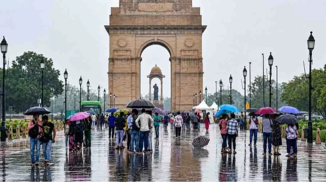 इंडिया गेट के पास बारिश के बीच छाता लेकर चलते लोग और आसमान में छाए बादल।