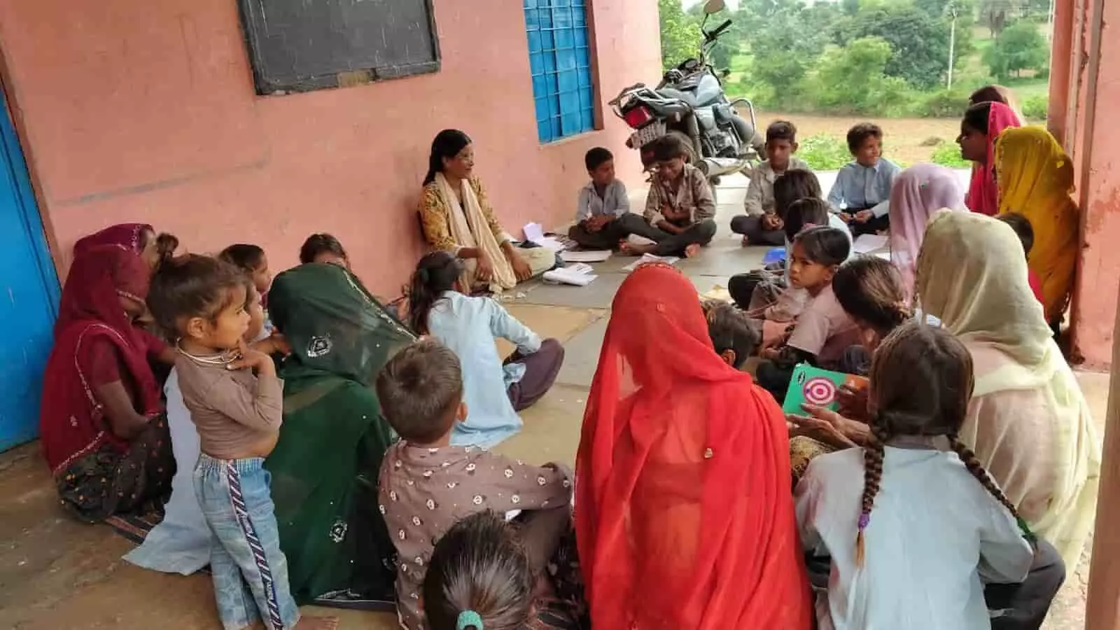 एक विद्यालय परिसर में बैठी महिलाएं और बच्चे जो लर्निंग वर्कशॉप में भाग ले रहे हैं।