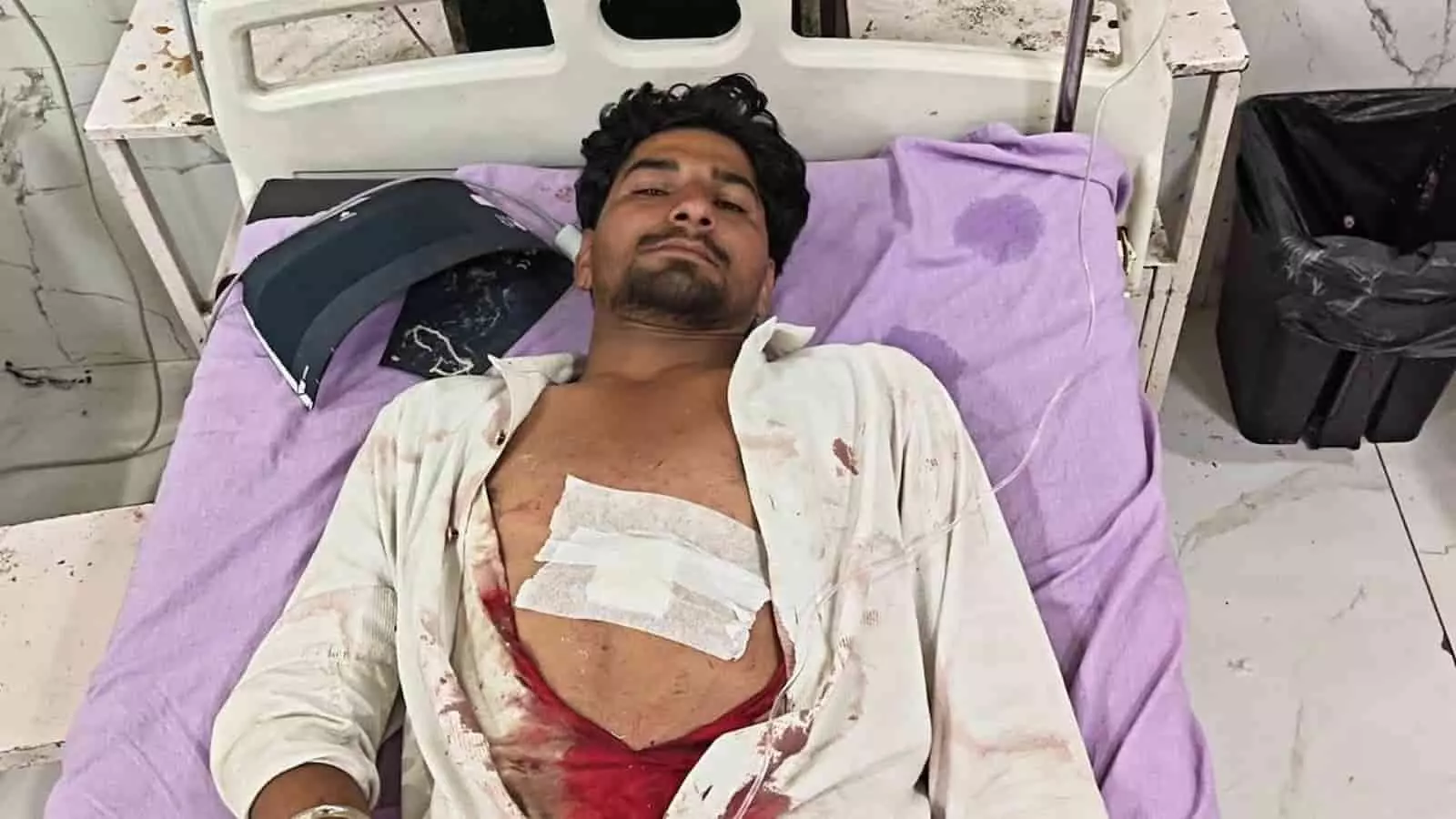 अस्पताल के बिस्तर पर लेटा हुआ घायल युवक जिसके सीने पर पट्टी बंधी है। अस्पताल के बिस्तर पर लेटा हुआ घायल युवक जिसके सीने पर पट्टी बंधी है।