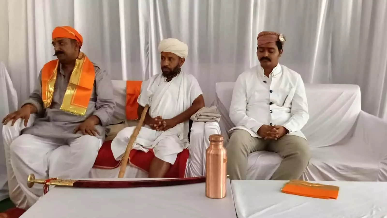मंच पर बैठे तीन अतिथियों के साथ कार्यक्रम में रखी एक तलवार और तांबे का पात्र। मंच पर बैठे तीन अतिथियों के साथ कार्यक्रम में रखी एक तलवार और तांबे का पात्र।