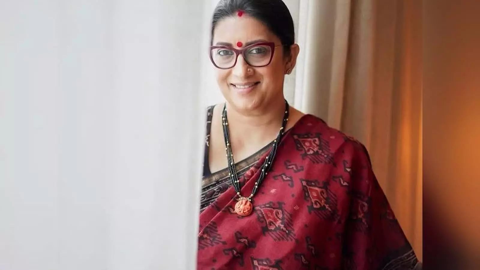 तस्वीर में मुस्कुराती हुई स्मृति ईरानी साड़ी पहने एक इनडोर पृष्ठभूमि के सामने खड़ी हैं। तस्वीर में मुस्कुराती हुई स्मृति ईरानी साड़ी पहने एक इनडोर पृष्ठभूमि के सामने खड़ी हैं।