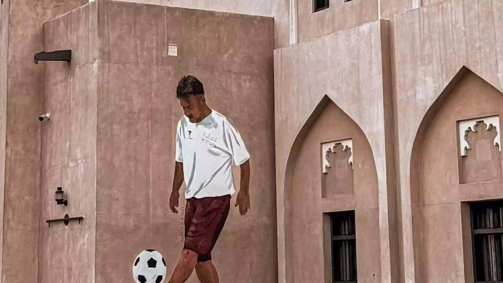 तस्वीर में कतर की पृष्ठभूमि पर बनी पारंपरिक अरब इमारतों के सामने फुटबॉल खेलता एक युवक दिख रहा है। तस्वीर में कतर की पृष्ठभूमि पर बनी पारंपरिक अरब इमारतों के सामने फुटबॉल खेलता एक युवक दिख रहा है।