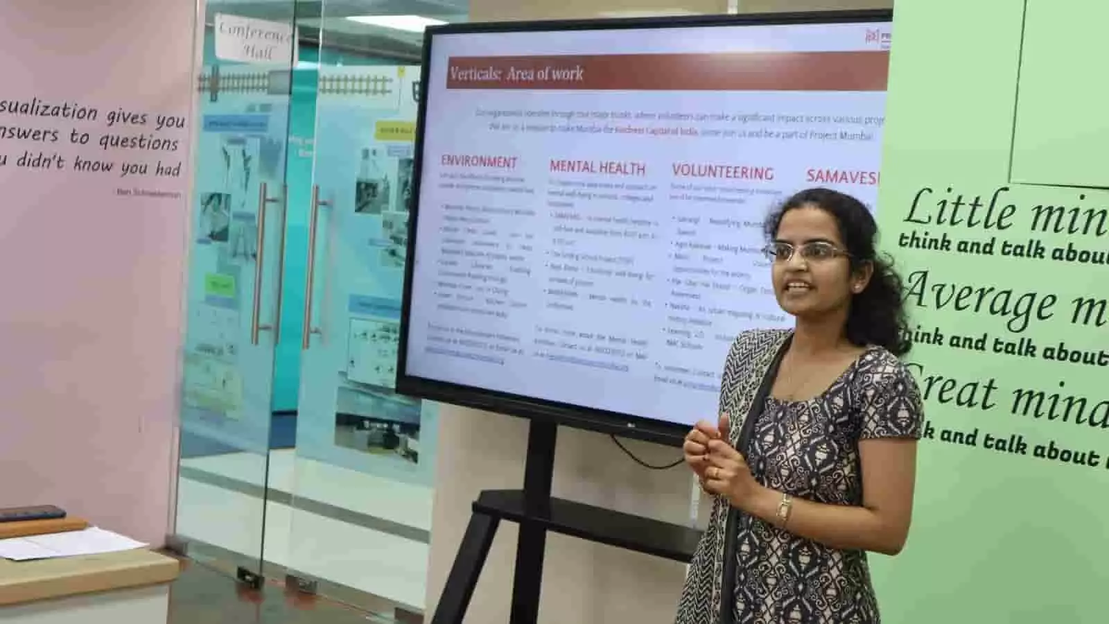 तस्वीर में एक महिला एक बड़ी स्क्रीन के सामने खड़ी होकर बात कर रही हैं। तस्वीर में एक महिला एक बड़ी स्क्रीन के सामने खड़ी होकर बात कर रही हैं।