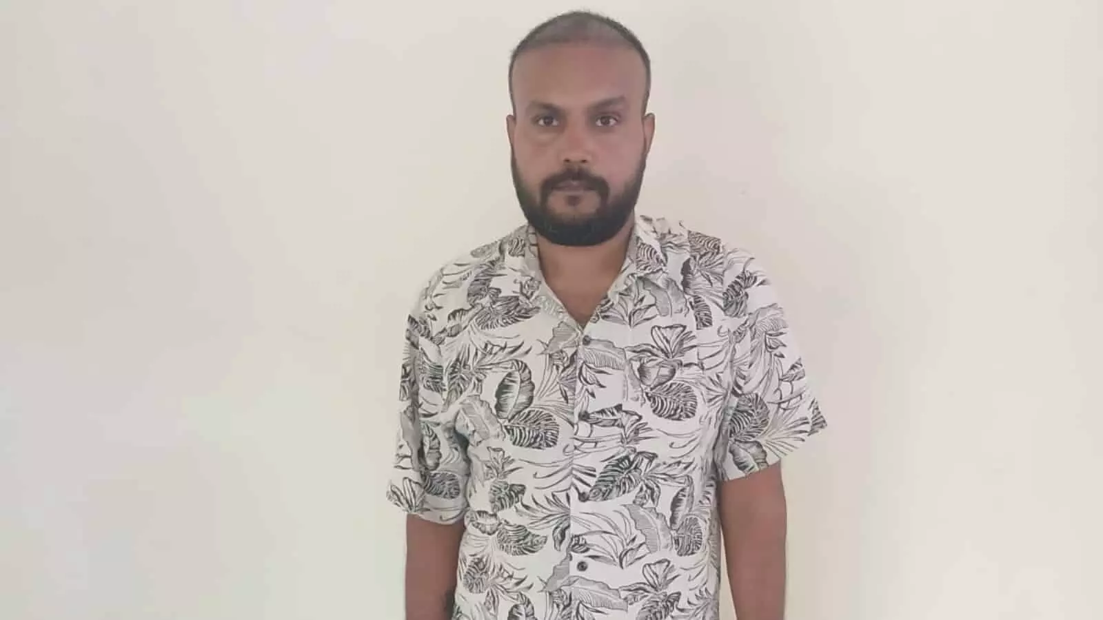 तस्वीर में सफेद प्रिंटेड शर्ट पहना एक दाढ़ी वाला पुरुष सादे बैकग्राउंड के सामने खड़ा है। तस्वीर में सफेद प्रिंटेड शर्ट पहना एक दाढ़ी वाला पुरुष सादे बैकग्राउंड के सामने खड़ा है।
