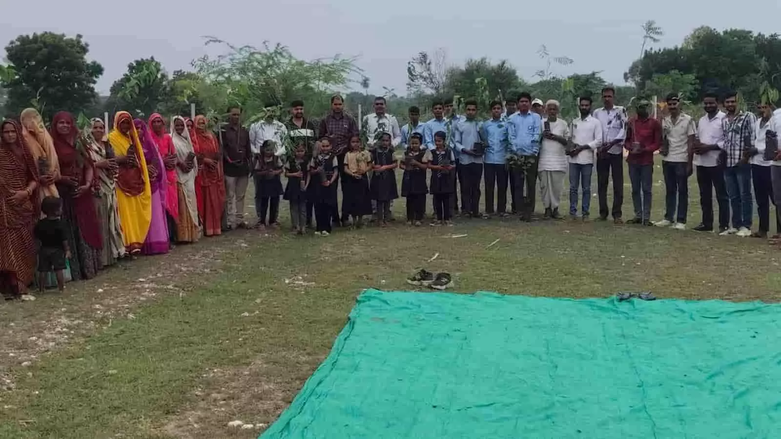 तस्वीर में खुले मैदान में हाथों में छोटे पौधे लेकर खड़े ग्रामीण और स्कूली बच्चे।