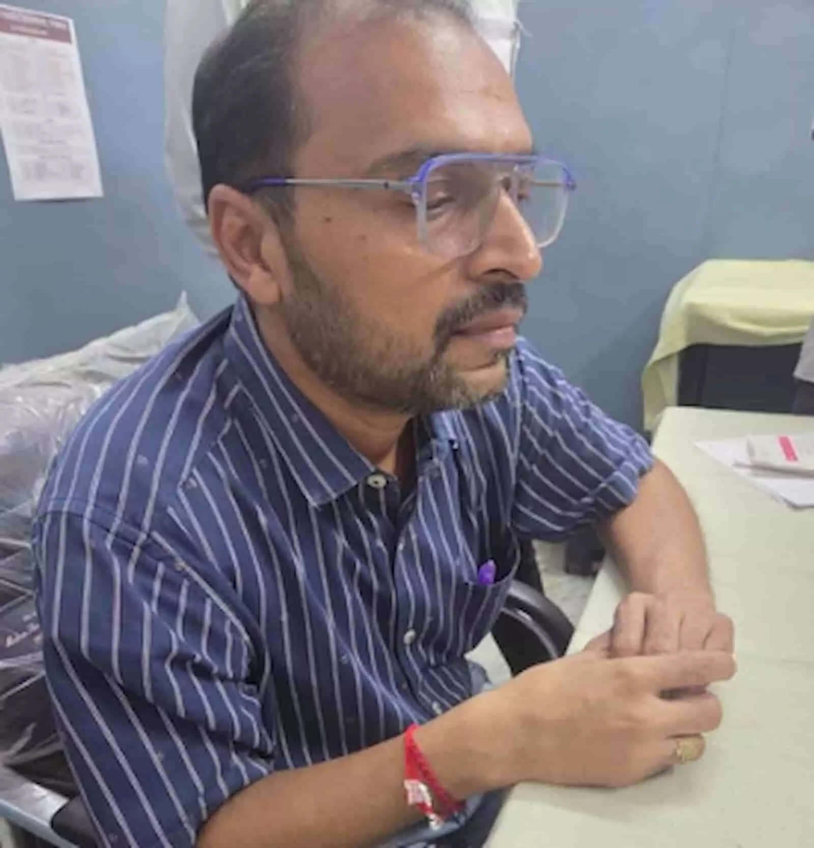 तस्वीर में कुर्सी पर बैठा चश्मा पहने एक व्यक्ति मेज पर हाथ रखे पूछताछ के संदर्भ में दिख रहा है। तस्वीर में कुर्सी पर बैठा चश्मा पहने एक व्यक्ति मेज पर हाथ रखे पूछताछ के संदर्भ में दिख रहा है।