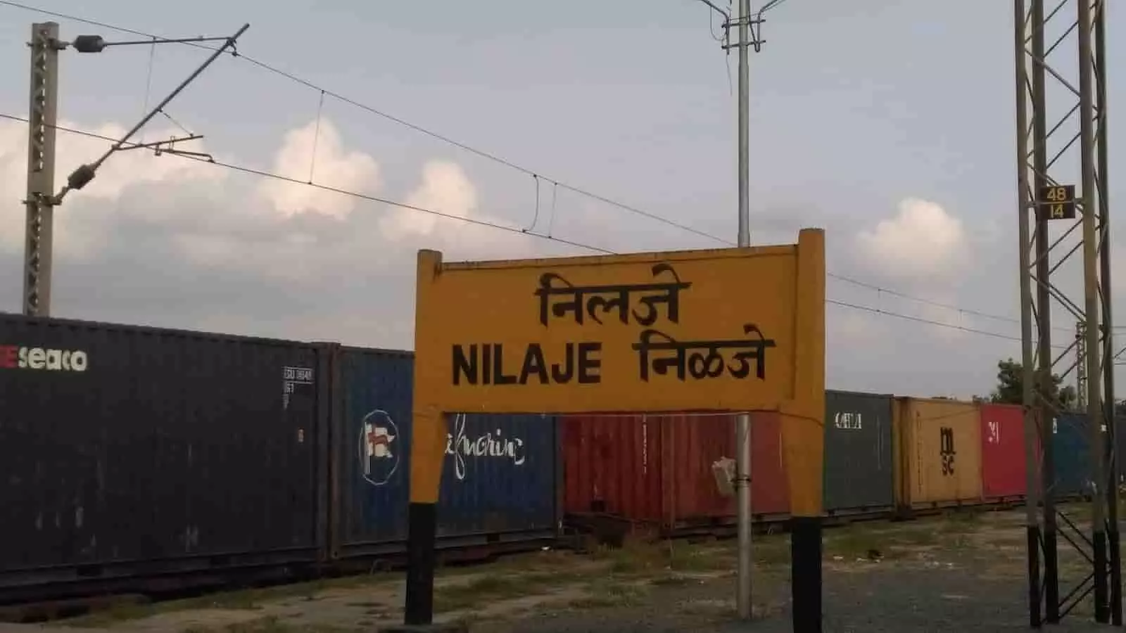 निलजे स्टेशन का पीला साइनबोर्ड और बैकग्राउंड में मालगाड़ी के कंटेनर दिख रहे हैं।