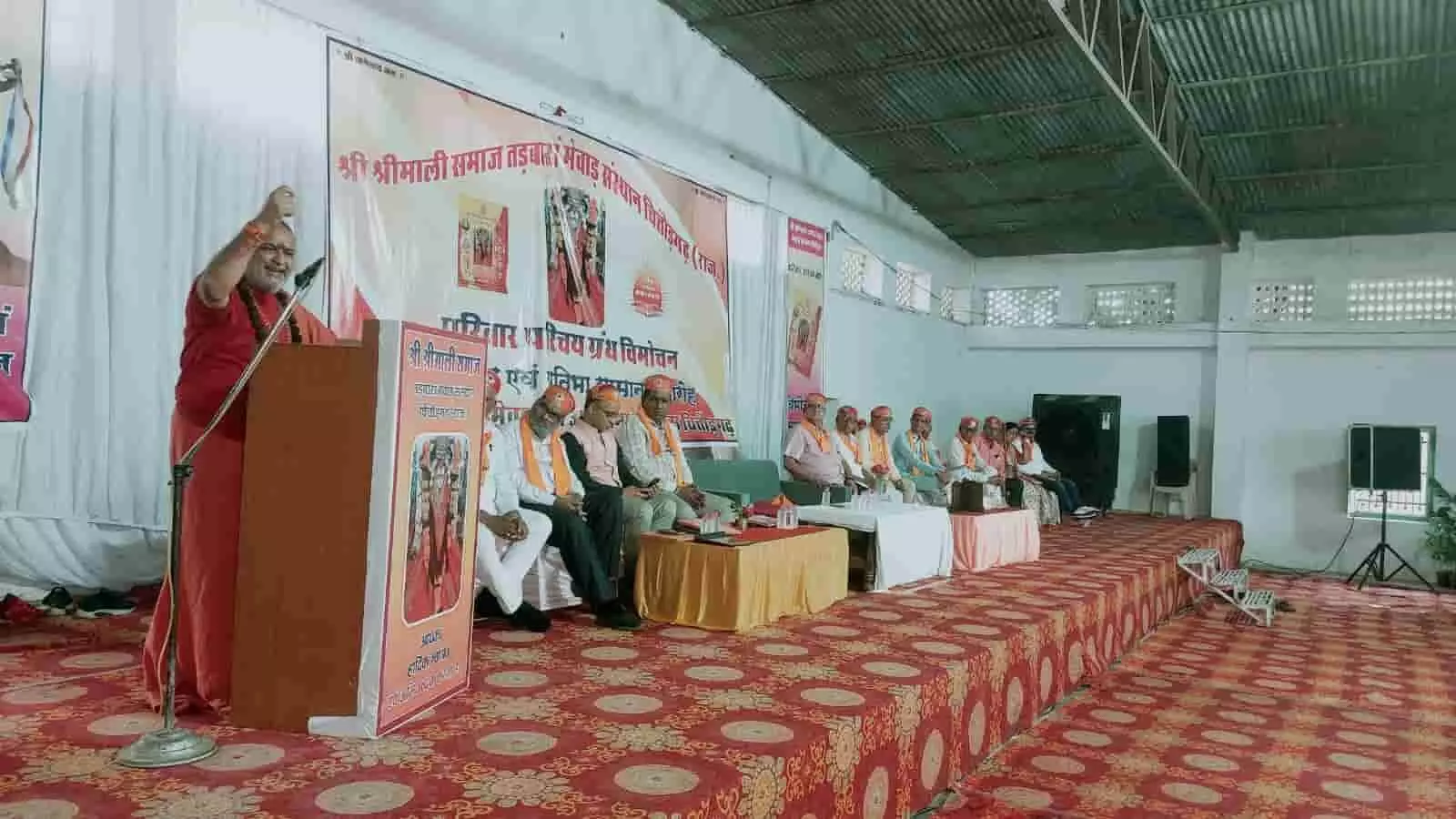 एक व्यक्ति मंच पर संबोधित कर रहे हैं और अतिथि सामने बैठे हैं। एक व्यक्ति मंच पर संबोधित कर रहे हैं और अतिथि सामने बैठे हैं।