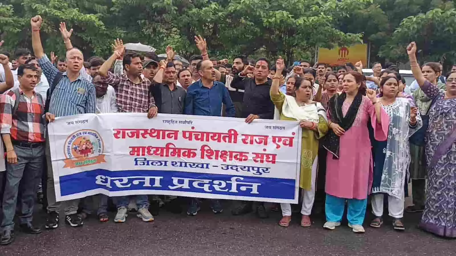 तस्वीर में उदयपुर में बैनर थामकर विरोध प्रदर्शन करते शिक्षक-शिक्षिकाएं दिख रहे हैं। तस्वीर में उदयपुर में बैनर थामकर विरोध प्रदर्शन करते शिक्षक-शिक्षिकाएं दिख रहे हैं।