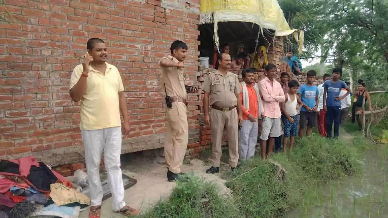 तस्वीर में ईंट के मकान के बाहर पुलिसकर्मी जांच करते और खड़े ग्रामीण दिख रहे हैं।
