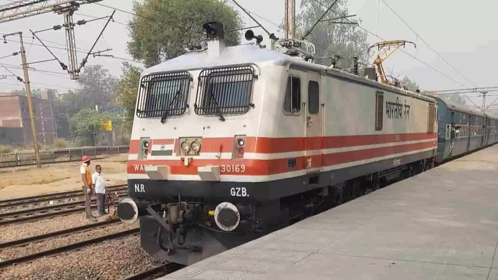 तस्वीर में रेलवे स्टेशन के प्लेटफॉर्म पर खड़ी एक ट्रेन और पटरियों के पास कर्मचारी दिख रहे हैं।