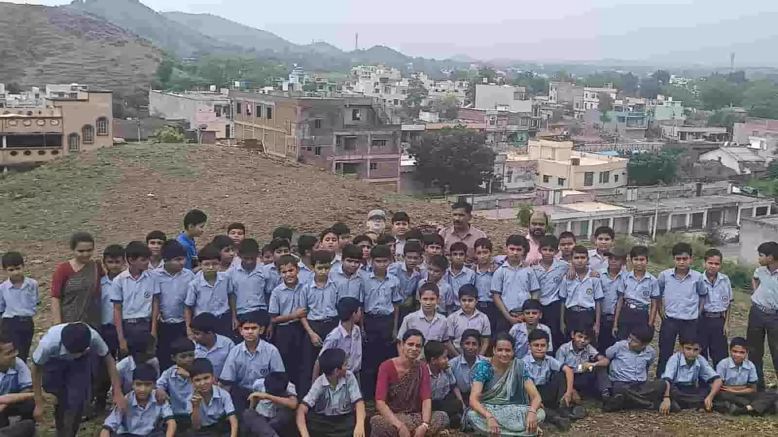 तस्वीर में पहाड़ी पर शिक्षकों के साथ सामूहिक रूप से बैठे एवं खड़े स्कूली बच्चे।