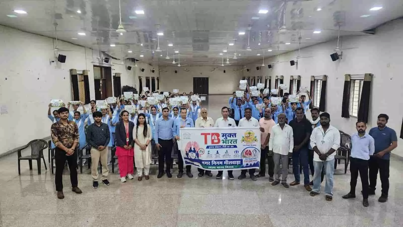 हॉल में टीबी मुक्त भारत का बैनर पकड़े स्वास्थ्यकर्मी और लोग खड़े दिख रहे हैं।