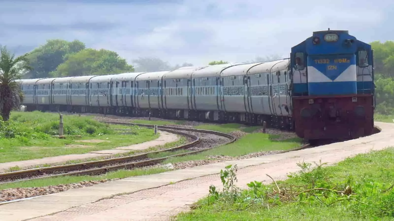 तस्वीर में नीले रंग का इंजन लगी एक पैसेंजर ट्रेन पटरी पर गुजरती हुई दिख रही है। तस्वीर में नीले रंग का इंजन लगी एक पैसेंजर ट्रेन पटरी पर गुजरती हुई दिख रही है।