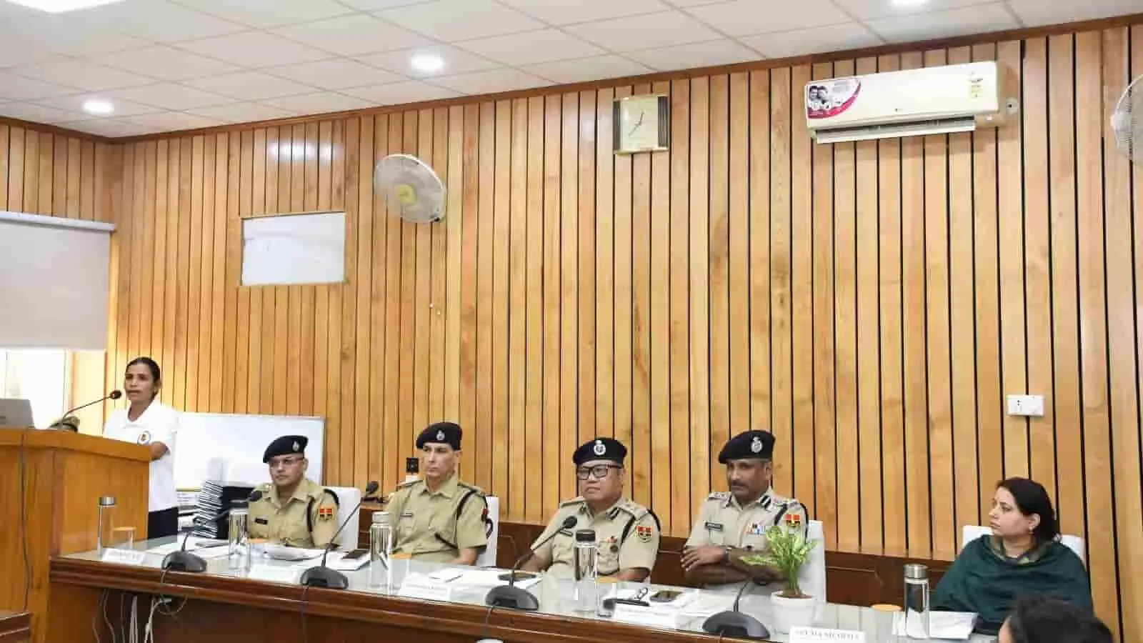 तस्वीर में मंच पर अधिकारी बैठे हैं और एक महिला पोडियम से सभा को संबोधित कर रही हैं। तस्वीर में मंच पर अधिकारी बैठे हैं और एक महिला पोडियम से सभा को संबोधित कर रही हैं।