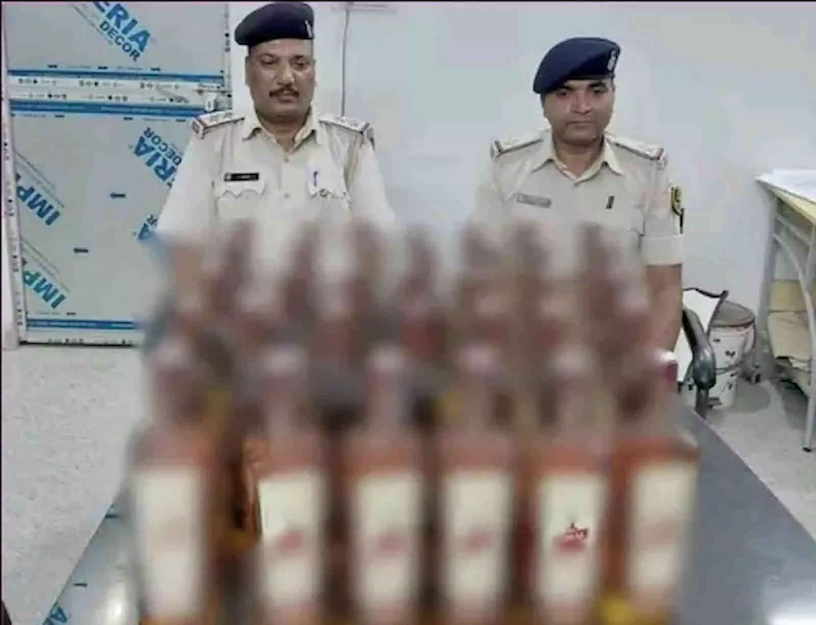 तस्वीर में दिख रही मेज पर रखी शराब की बोतलों के पीछे दो पुलिस अधिकारी खड़े हैं। तस्वीर में दिख रही मेज पर रखी शराब की बोतलों के पीछे दो पुलिस अधिकारी खड़े हैं।