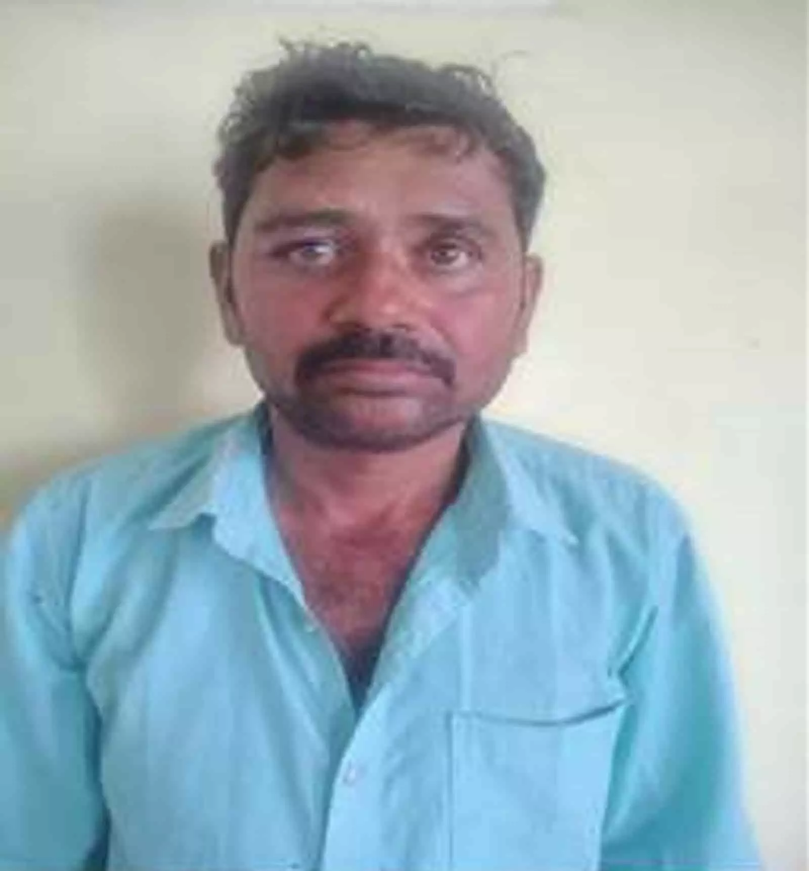 तस्वीर में दीवार के सामने नीली शर्ट पहने खड़ा गिरफ्तार आरोपी दिख रहा है।