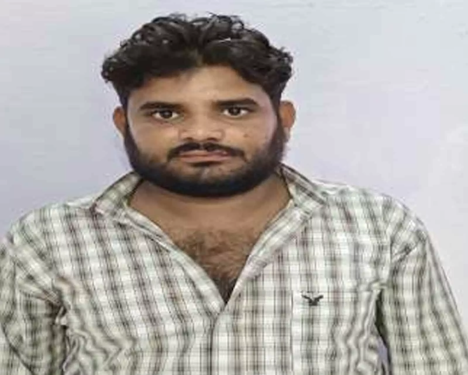 तस्वीर में चेक्ड शर्ट पहने एक दाढ़ी वाला व्यक्ति साधारण पृष्ठभूमि के सामने खड़ा दिख रहा है।