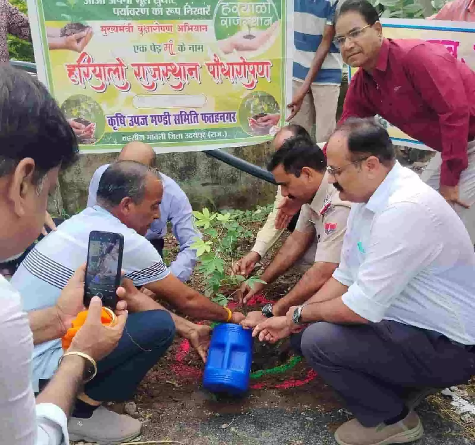 तस्वीर में हरियालो राजस्थान बैनर के सामने मिट्टी में पौधा लगाते और पानी देते लोग। तस्वीर में हरियालो राजस्थान बैनर के सामने मिट्टी में पौधा लगाते और पानी देते लोग।