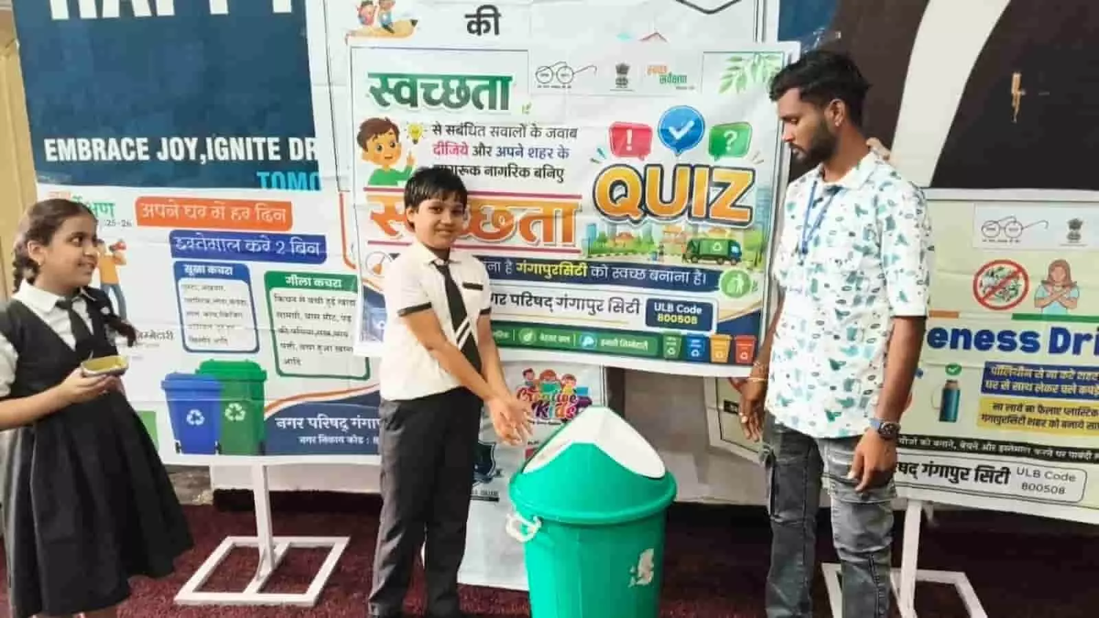 तस्वीर में एक छात्र विद्यालय में हरे रंग के डस्टबिन के पास खड़ा है। तस्वीर में एक छात्र विद्यालय में हरे रंग के डस्टबिन के पास खड़ा है।