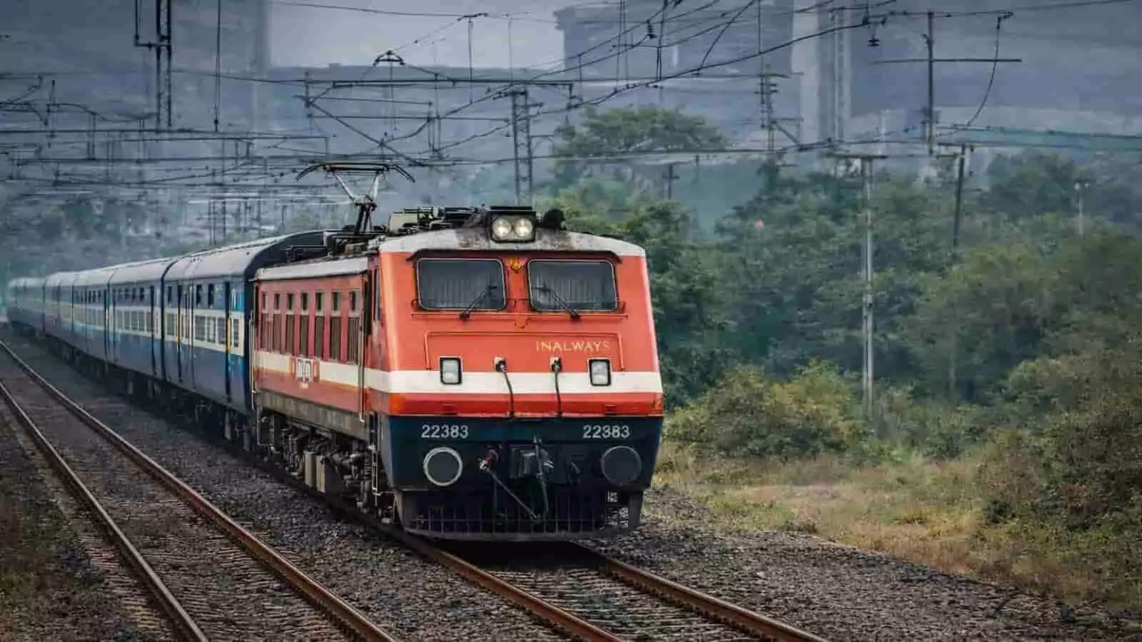 पटरी पर चलती रेलगाड़ी का लाल-सफेद इलेक्ट्रिक इंजन, पीछे नीली बोगियां, आसपास पेड़ और बिजली के तार। पटरी पर चलती रेलगाड़ी का लाल-सफेद इलेक्ट्रिक इंजन, पीछे नीली बोगियां, आसपास पेड़ और बिजली के तार।
