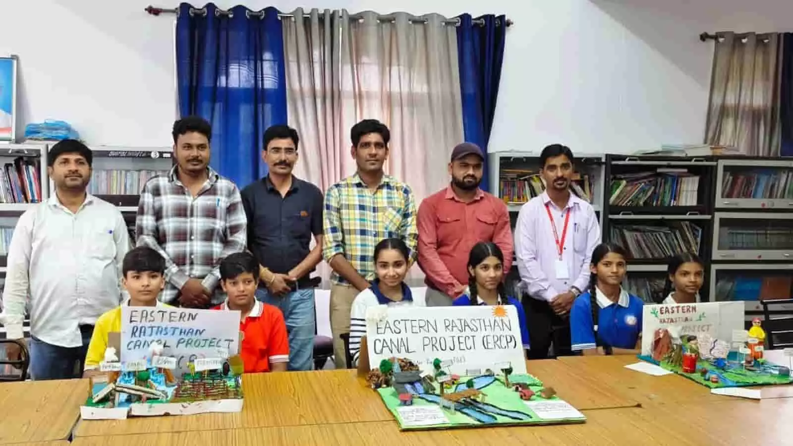 विद्यालय कक्ष में मेज पर रखे मॉडलों के साथ खड़े और बैठे छात्र व शिक्षक। विद्यालय कक्ष में मेज पर रखे मॉडलों के साथ खड़े और बैठे छात्र व शिक्षक।