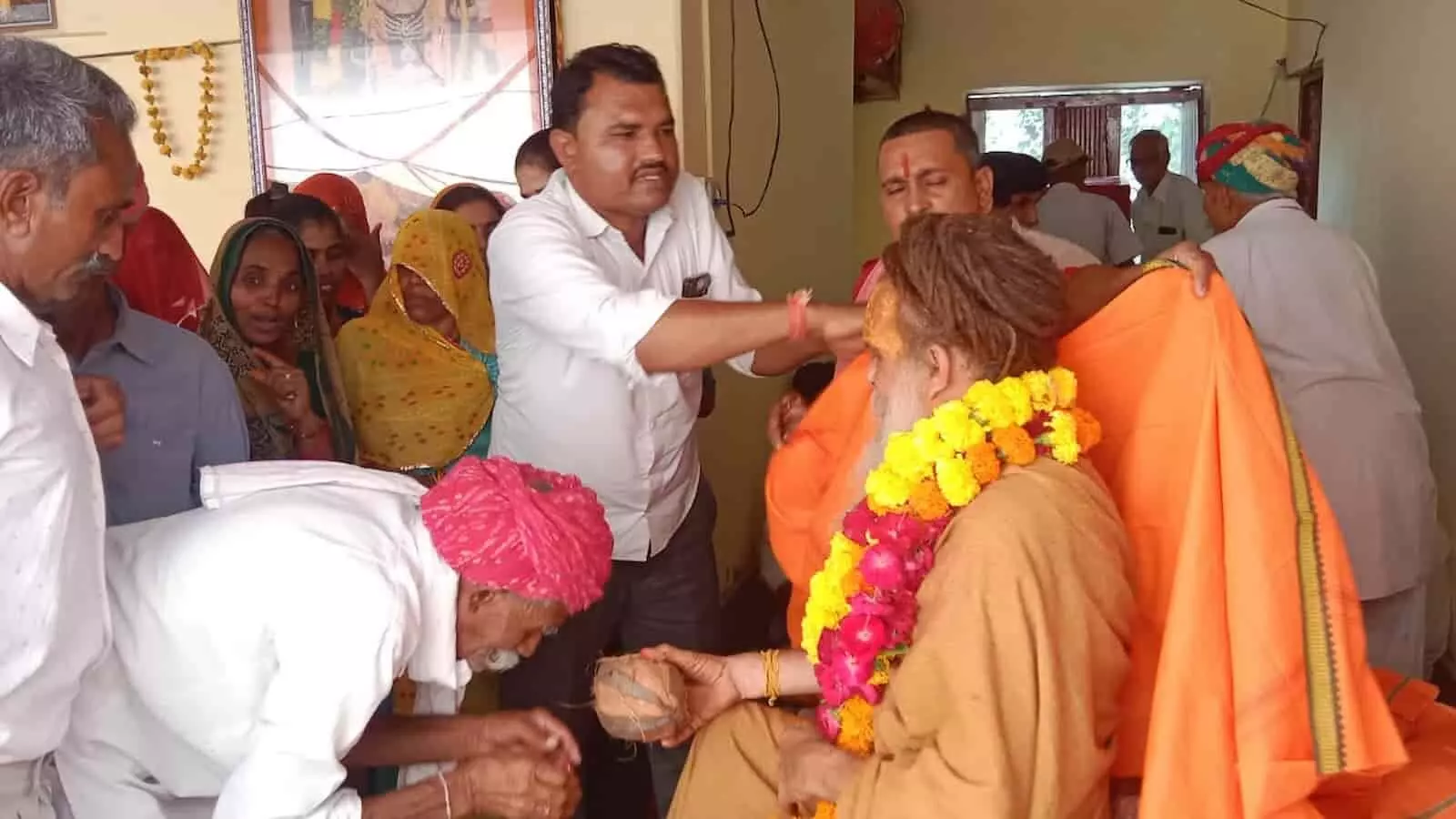 तस्वीर में एक मंदिर परिसर में संत को शॉल ओढ़ाते और आशीर्वाद लेते हुए श्रद्धालु दिख रहे हैं।