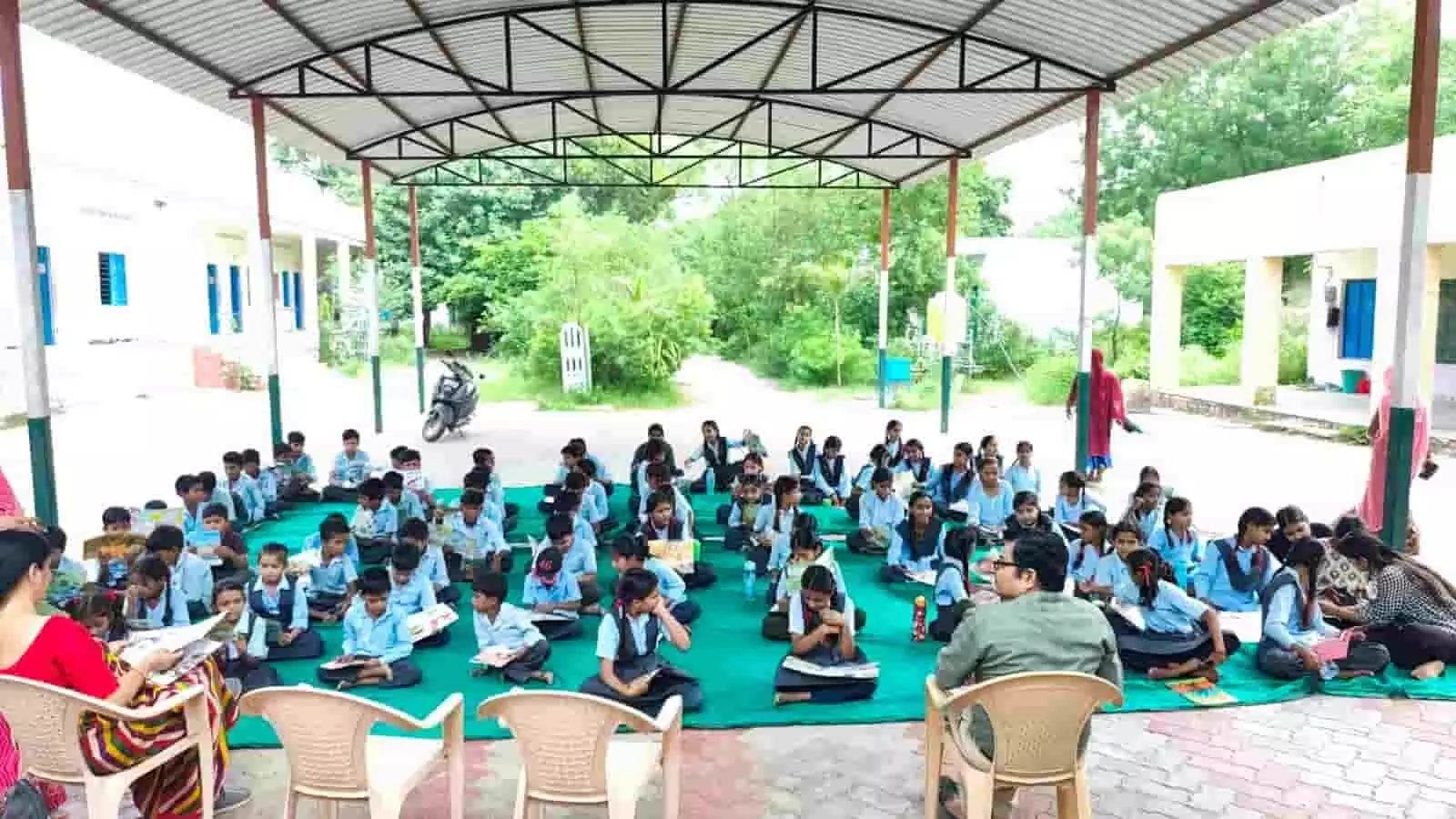 विद्यालय परिसर में जमीन पर बैठकर किताबें पढ़ते छात्र और कुर्सी पर बैठे शिक्षक। विद्यालय परिसर में जमीन पर बैठकर किताबें पढ़ते छात्र और कुर्सी पर बैठे शिक्षक।