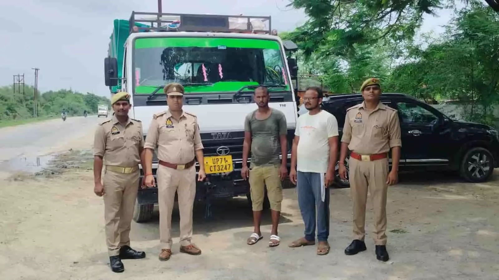 तस्वीर में पुलिसकर्मी और दो आरोपी खड़े हैं, पीछे सफेद डीसीएम वाहन खड़ा दिखाई दे रहा है।