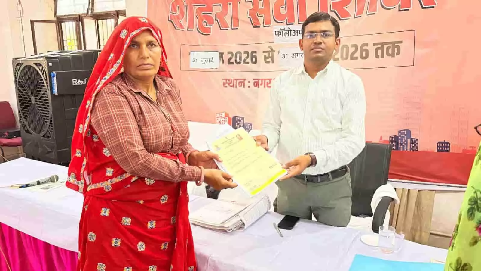 तस्वीर में एक महिला शहरी सेवा शिविर के दौरान अधिकारी से दस्तावेज लेते हुए दिख रही है। तस्वीर में एक महिला शहरी सेवा शिविर के दौरान अधिकारी से दस्तावेज लेते हुए दिख रही है।