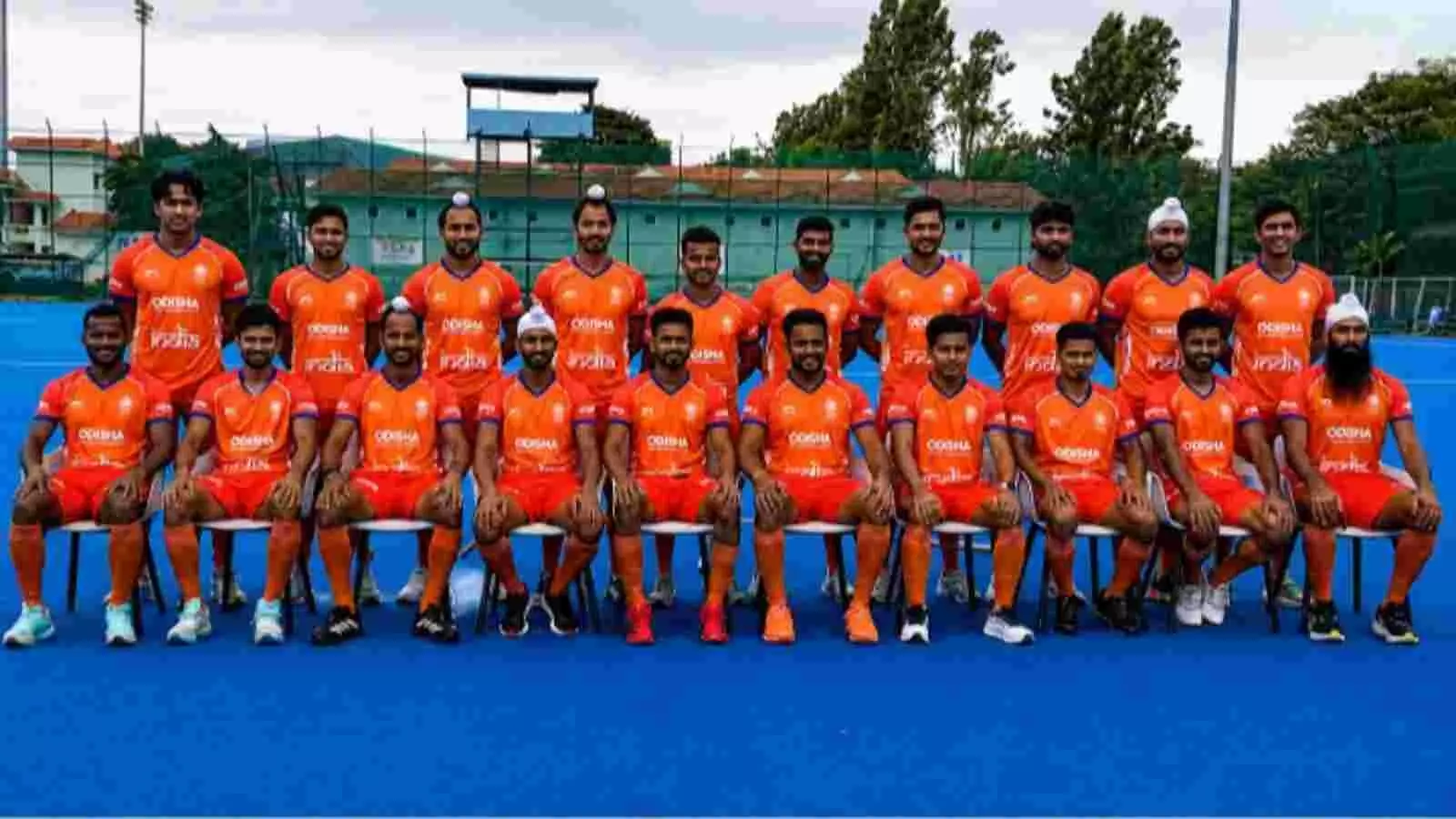 केसरिया जर्सी पहने भारतीय हॉकी टीम के खिलाड़ी नीले टर्फ मैदान पर खड़े और बैठे हैं। केसरिया जर्सी पहने भारतीय हॉकी टीम के खिलाड़ी नीले टर्फ मैदान पर खड़े और बैठे हैं।